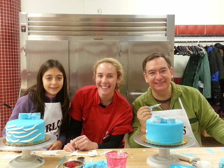 A smiling man, woman, and girl with two blue layer cakes and white fondant icing.