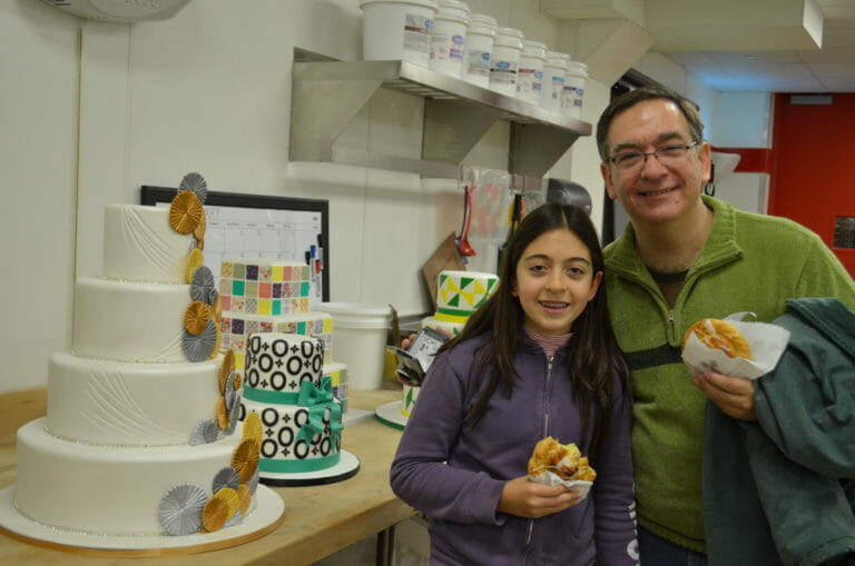 A man in glasses and a girl holding sweet rolls in a bakery, by layered cakes decorated with fondant icing.