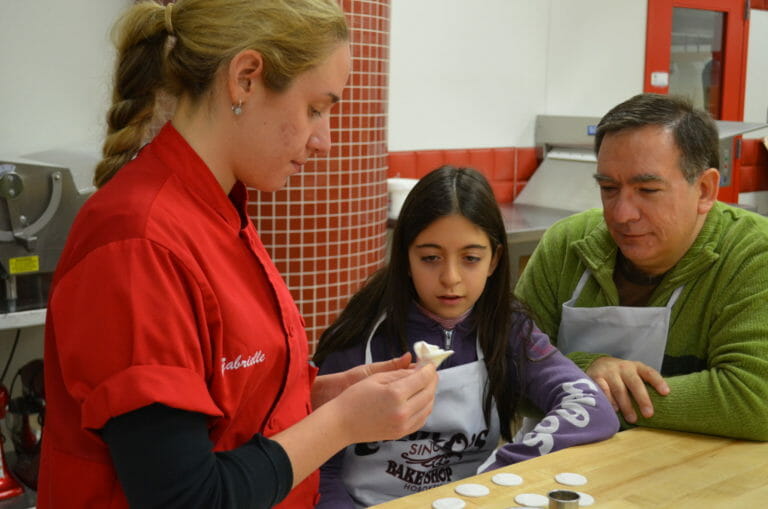 A bakery employee in a red smock showing a man and a girl how to make fondant roses.
