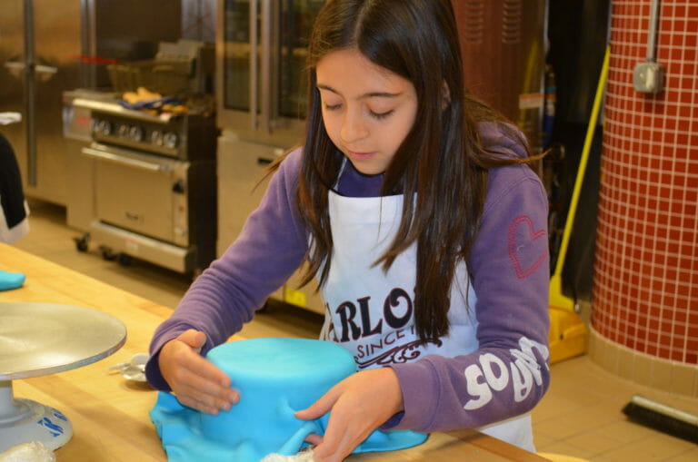 A girl in a Carlo's Bake Shop apron wrapping blue fondant over a layer cake.
