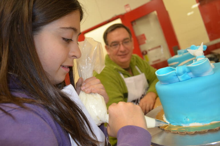 A man in glasses watches as a girl in a bakery squeezes white icing around the bottom of a blue layer cake.
