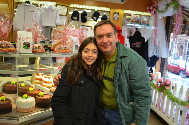 A man and a girl hugging in a bakery, surrounded by cakes, cookies, and other bakery items.