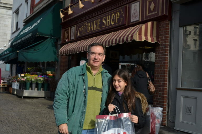 A man and a girl outside Carlo's Bake Shop, holding a shopping bag of goodies.