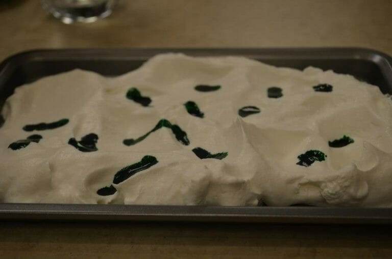 A baking pan filled with whipped topping, with green food color puddles on top.