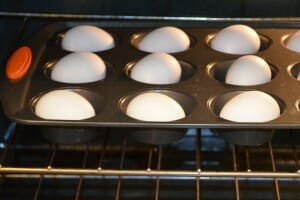 A dozen white eggs in shells placed in a muffin pan that is on a rack in an oven.