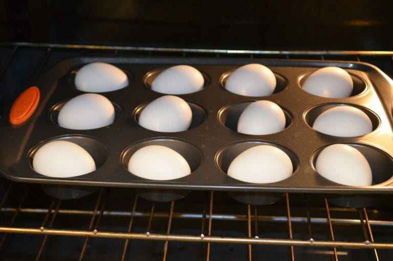 A dozen white eggs in shells placed in a muffin pan that is on a rack in an oven.