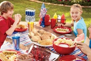A boy and a girl at a picnic table decorated in red, white, and blue, and filled with barbeque party food.