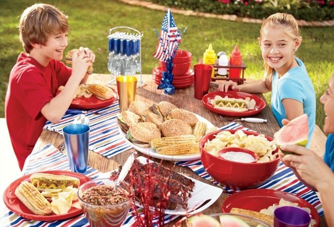 A boy and a girl at a picnic table decorated in red, white, and blue, and filled with barbeque party food.