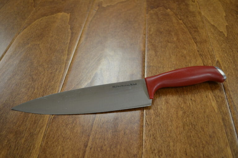 A KitchenAid Chef's Knife with a red handle placed on a wood table.