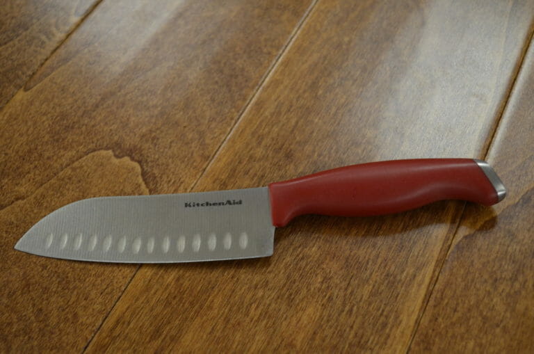A KitchenAid Santoku Knife with a red handle placed on a wood table.