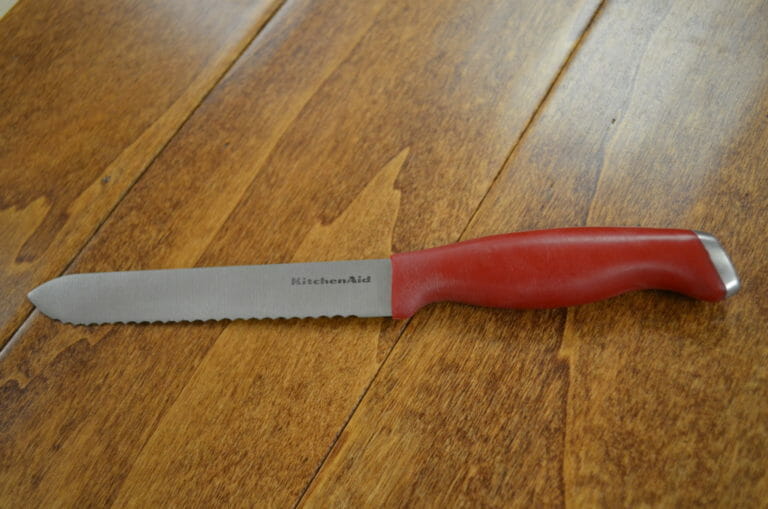 A KitchenAid Serrated Utility Knife with a red handle placed on a wood table.