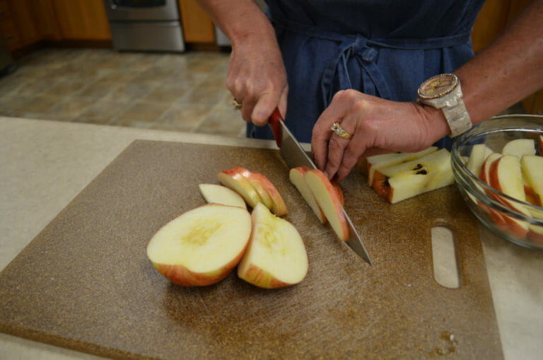 A woman cutting apple slices on a brown cutting board with a red-handled knife.