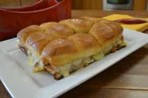 A white platter filled with 12 baked ham and Swiss cheese sandwiches on Hawaiian rolls.
