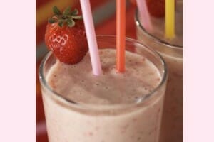 A strawberry shake in a clear glass with two straws and a fresh strawberry garnish,