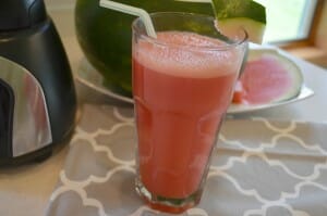 A clear glass with a straw, filled with pink Melon Refresher, next to a cut watermelon.