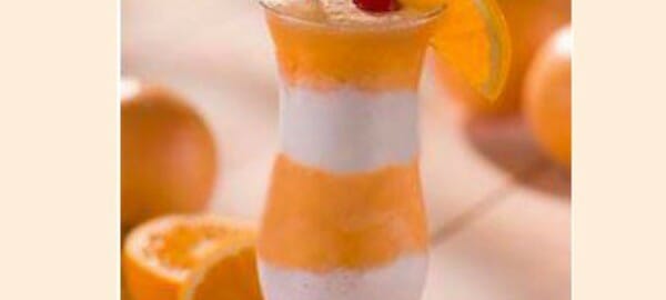 A clear curved glass filled with layered orange and pink Orange Cooler, with a straw, an orange slice and a cherry.