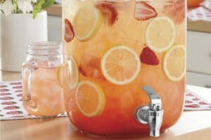 A clear mug and a drink dispenser filled with strawberry iced tea, with floating orange and strawberry slices.