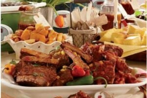 A buffet table filled with a platter of Braised BBQ Short Ribs, cornbread, home fries, plastic flatware, and salad.