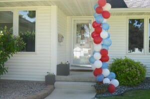 Red, white, and blue balloons wrapped around a porch post by the front door of a white house.