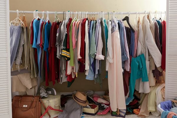 Interior of a messy closet with just one rod for hanging clothes, and piles of shoes, purses, and more on the floor.