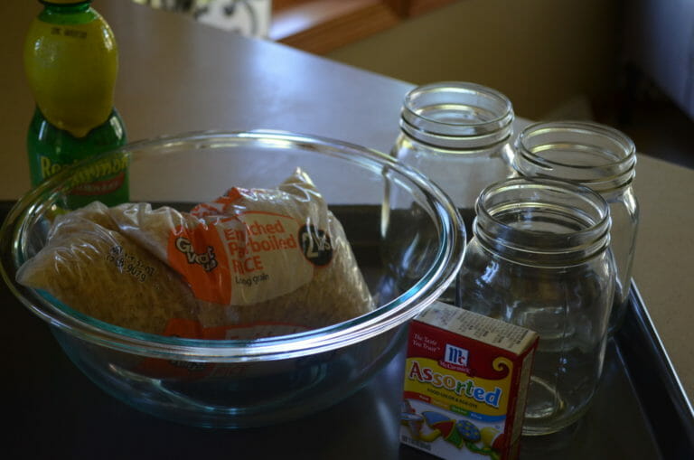 Three empty Mason jars, a box of food coloring, a bottle of ReaLemon, and a clear mixing bowl holding a bag of rice.