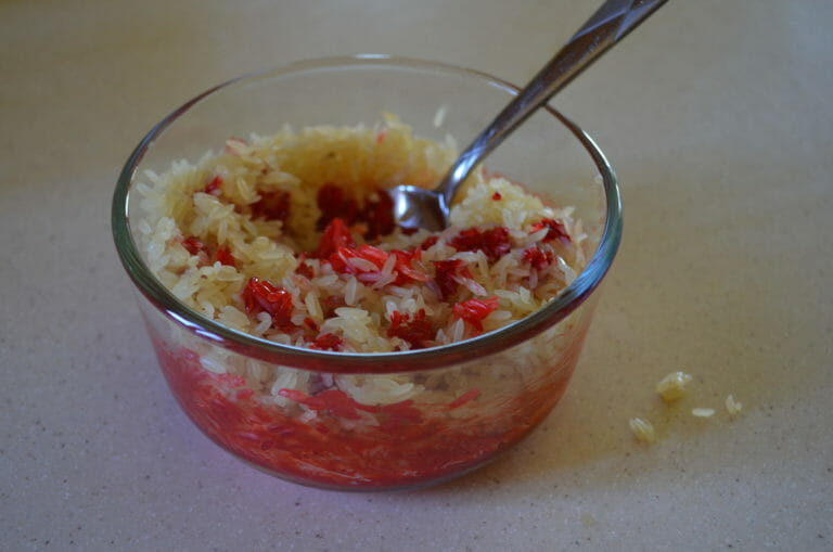 A spoon in a clear mixing bowl with parboiled rice and red food coloring.