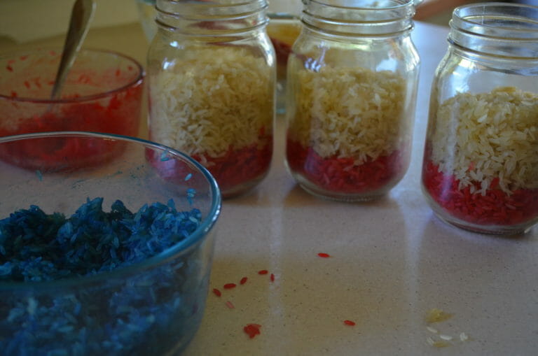 Three Mason jars filled with red rice on the bottom, white rice in the middle, and a bowl of blue rice nearby.