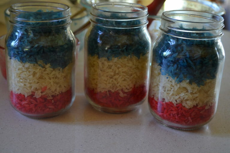 Three Mason jars filled with red rice on the bottom, white rice in the middle, and blue rice on top.