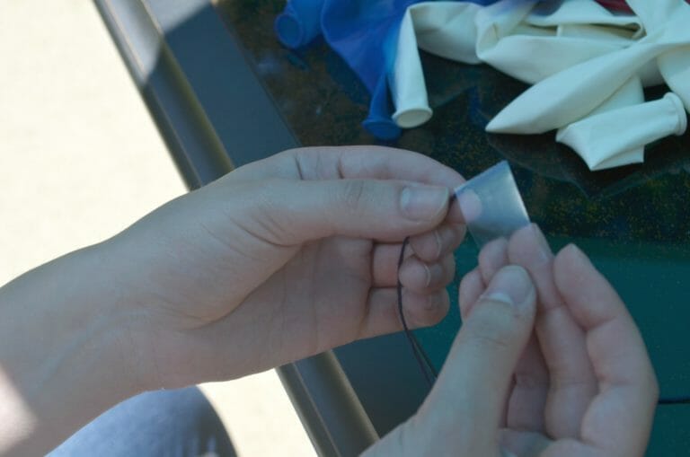 A person's hands wrapping tape around an end of blue embroidery floss, near a pile of deflated balloons.