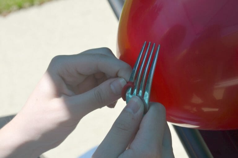 A person's hands poking a hole into the end of a blown up balloon with the tine of a fork.