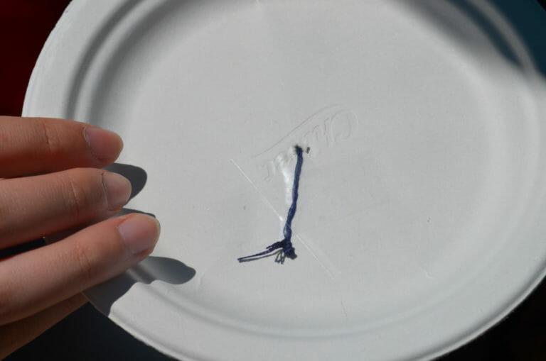 A white Chinet paper plate with a strand of blue embroidery floss poked through the center, and taped down.