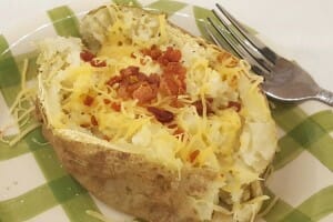 A baked potato with bacon bits and shredded cheese on a green check plate with a fork.