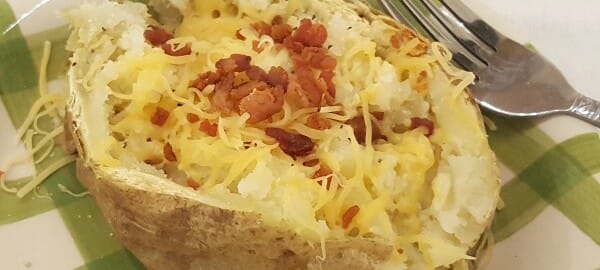 A baked potato with bacon bits and shredded cheese on a green check plate with a fork.