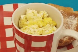 A red and white check mug filled with microwaved eggs, by a buttered slice of toast.