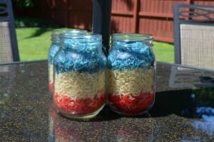 Three Mason jars on a patio table, filled with layers of red, white, and blue parboiled rice.