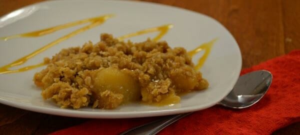 A serving of Apple Crisp with drizzles of caramel on a white plate, with a spoon on a red napkin.