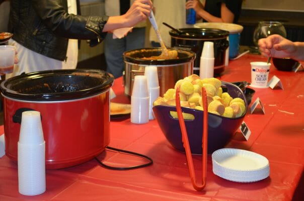 A long table covered in red plastic, with four slow cookers lined up for a Chili Cookoff, with a bowl of cornbread muffins.