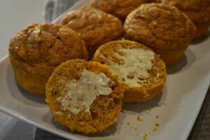 A white platter filled with pumpkin muffins, with one sliced open and spread with butter.