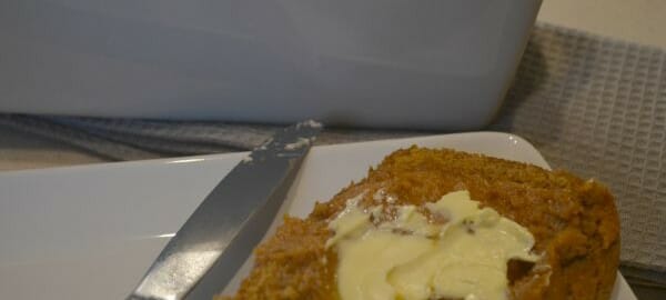 A slice of pumpkin bread on a white plate, spread with butter by a nearby knife.