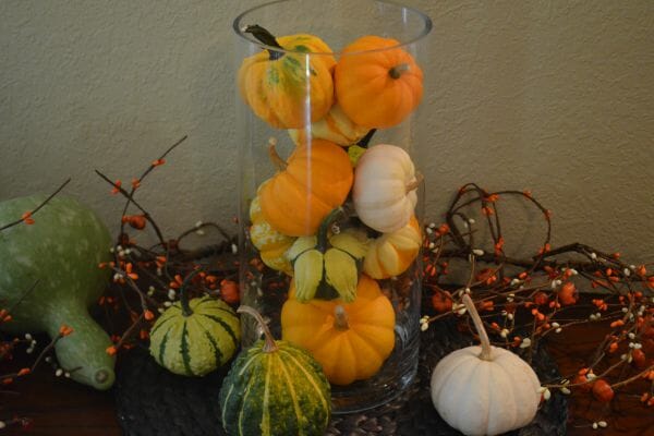 A clear glass vase on a mantel filled with mini gourds, and surrounded by other gourds and silk bittersweet vines.