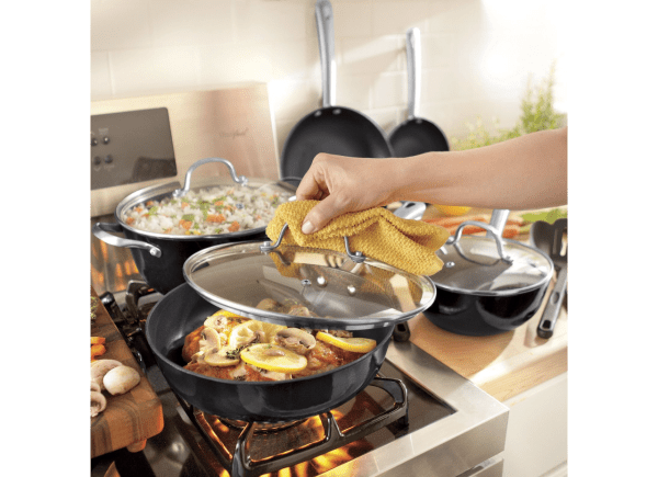 Woman lifting a glass lid from a pan filled with lemon chicken, while a kettle with vegetable rice cooks on the gas stove.
