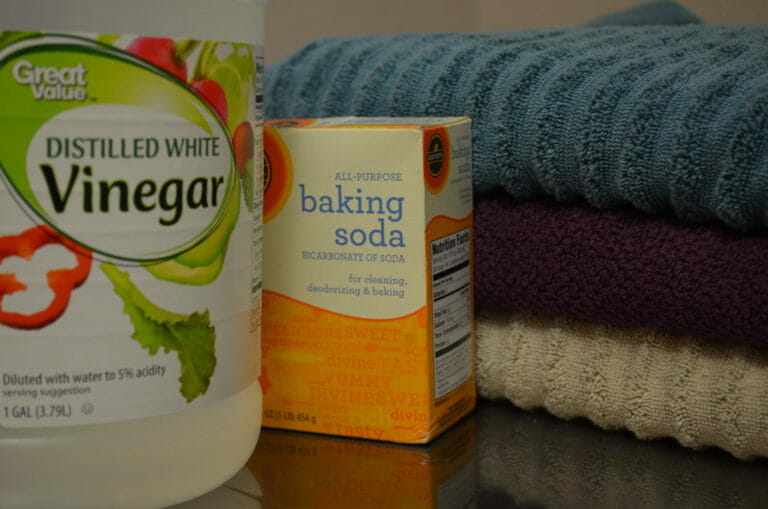 A bottle of white vinegar, a box of baking soda, and three folded towels in blue, plum, and tan.