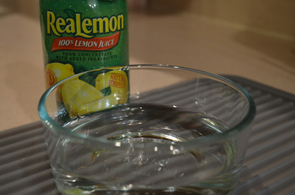 A bottle of ReaLemon and a clear bowl of water on a gray mat.
