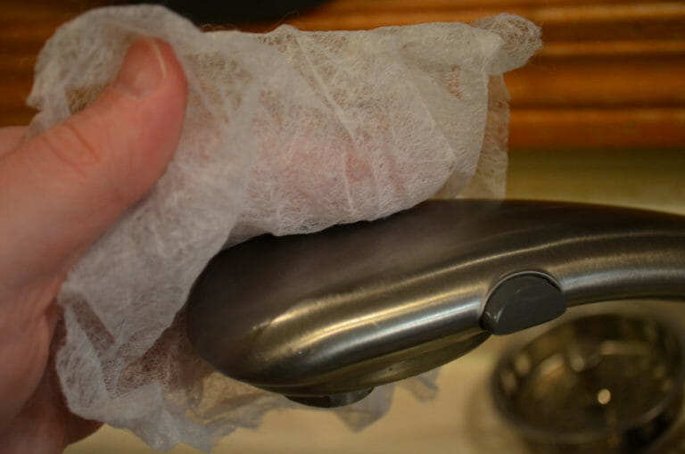 Close-up of a person wiping a brushed metal sink faucet with a dryer sheet.