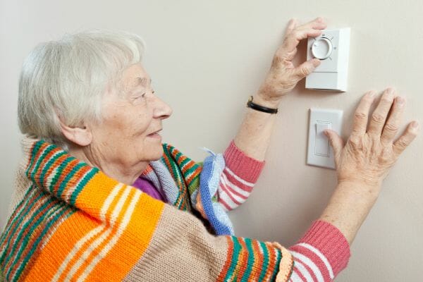 An elderly woman wrapped in a knit blanket, adjusting a wall thermostat.