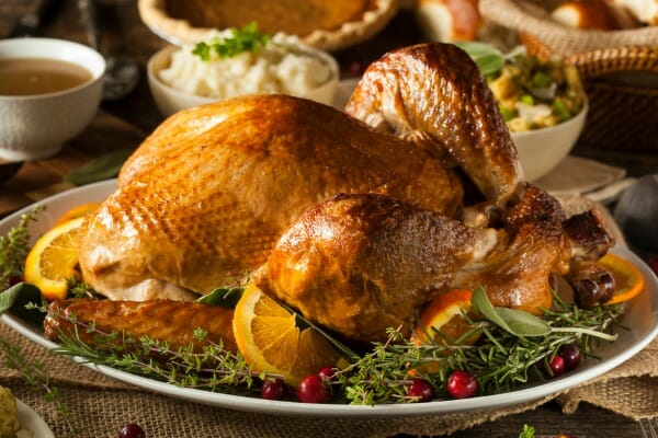 A roast turkey on a platter with herbal garnishes, with bowls of mashed potatoes, gravy, rolls, and pie on a table.