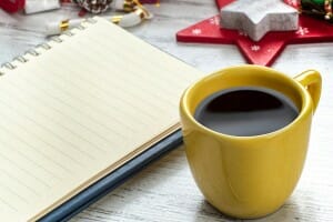 A yellow cup of coffee next to a blank lined notebook, on a table with Christmas decorations.