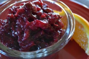 A clear bowl with Cranberry Orange Sauce, on a red plate with an orange slice.
