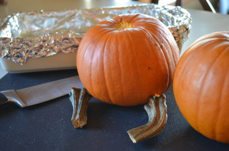 Two small pumpkins on a cutting board, with the stems removed, next to a knife and a foil lined pan.