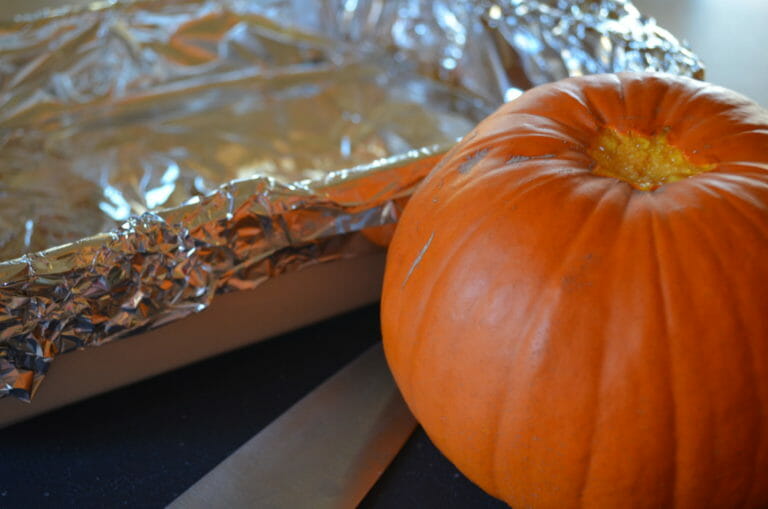 A small pumpkin on a cutting board, with the stem removed, next to a knife and a foil lined pan.
